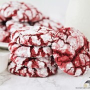 a stack of red velvet cake mix cookies with a dusting of powdered sugar next to a glass jar of milk. Behind these are another plate full of more cookies.