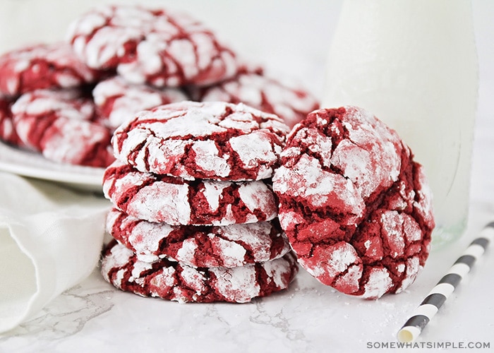 a stack of red velvet cake mix cookies with a dusting of powdered sugar next to a glass jar of milk. Behind these are another plate full of more cookies.