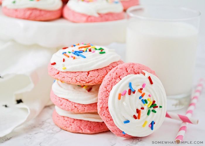 a stack of pink strawberry cookies topped with a white cream cheese frosting and colored sprinkles