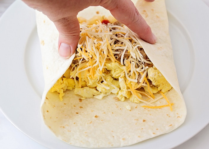 a hand folding over the edges of a tortilla filled with ingredients to make a breakfast burrito