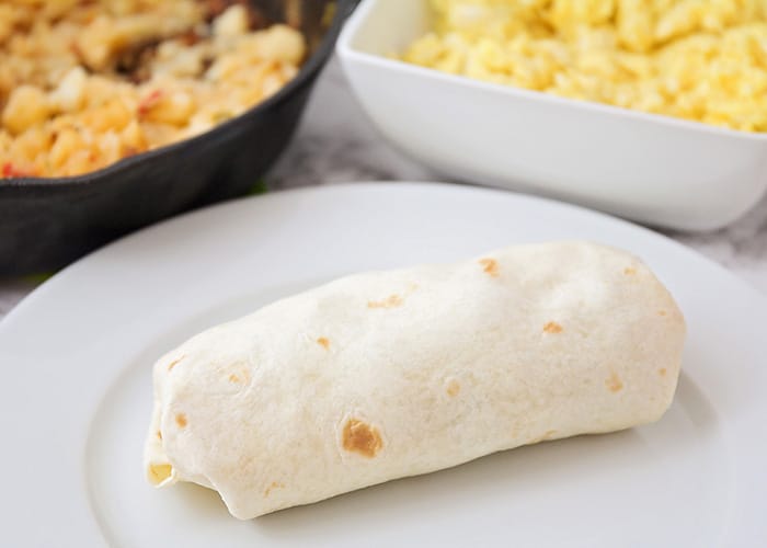 a burrito on a plate with eggs a bowl of eggs and a skillet of breakfast potatoes in the background