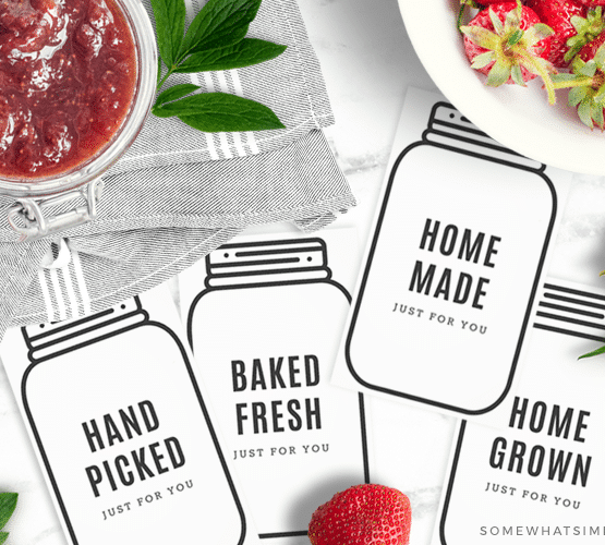 several mason jar gift tags cut out and laid on the counter next to fresh strawberries and a jar of strawberry jam