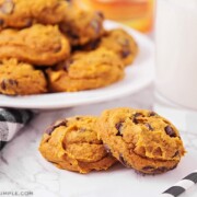 two pumpkin cake mix chocolate chip cookies on a counter next to a glass of milk and a plate of cookies in the background