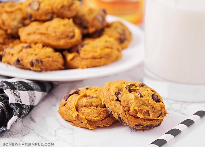 two pumpkin cake mix chocolate chip cookies on a counter next to a glass of milk and a plate of cookies in the background