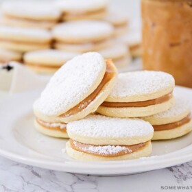 a plate of homemade alfajores with powdered sugar on a table made using this easy recipe with a jar of dulce de leche behind it