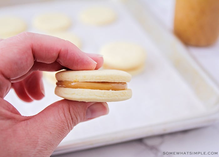 una mano sosteniendo un alfajore con dulce de leche marrón claro dentro de la galleta