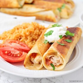 two crispy chicken taquitos on a plate topped with sour cream and a side of rice
