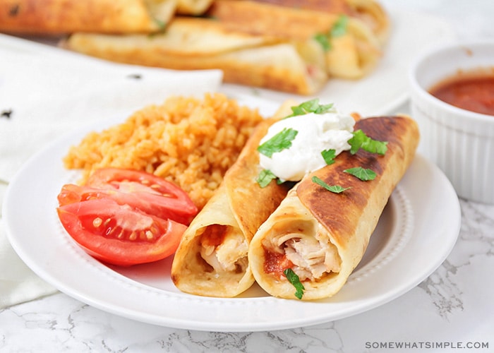 two crispy chicken taquitos on a plate topped with sour cream and a side of rice