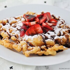 a homemade funnel cake on a white plate topped with freshly sliced strawberries, powdered sugar and chocolate syrup