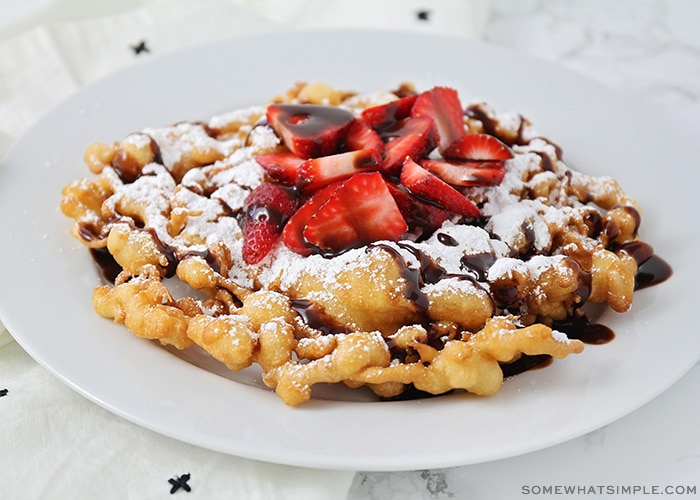 a homemade funnel cake on a white plate topped with freshly sliced strawberries, powdered sugar and chocolate syrup