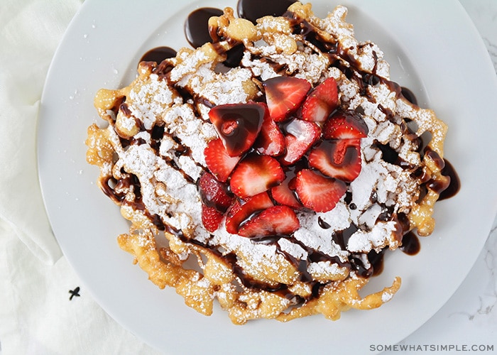 looking down on a funnel cake topped with powdered sugar, strawberries and chocolate syrup