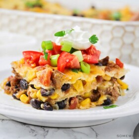 a square of taco casserole filled with ground beef, cheese, beans and corn and topped with diced tomatoes and green onions with a dollop of sour cream
