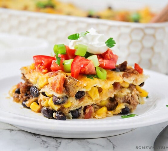 a square of taco casserole filled with ground beef, cheese, beans and corn and topped with diced tomatoes and green onions with a dollop of sour cream