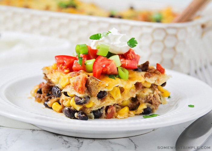 a square of taco casserole filled with ground beef, cheese, beans and corn and topped with diced tomatoes and green onions with a dollop of sour cream 