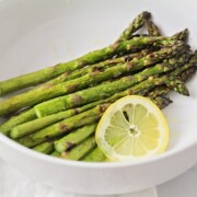 stalks of grilled asparagus in a white bowl next to a lemon slice