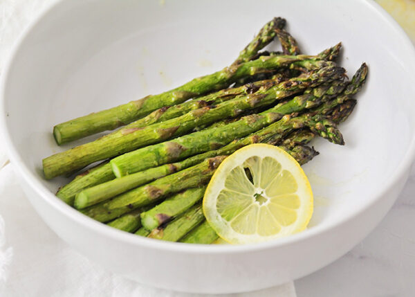 stalks of grilled asparagus in a white bowl next to a lemon slice