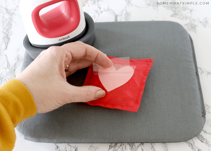 red bean bag with pink heart on a gray mat and hand pulling off plastic sheet from the iron-on material. Small iron in the back corner. 