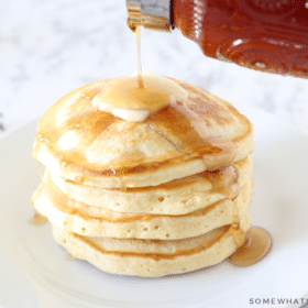 pouring syrup on top of a stack of pancakes with a slab of butter
