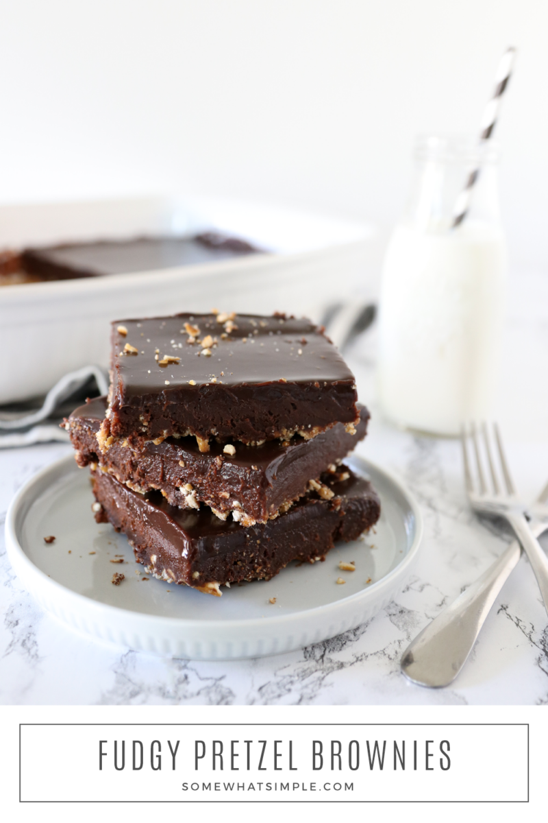 three stacked pretzel crusted brownies on a white plate with the pan of brownies and a glass of milk in the background