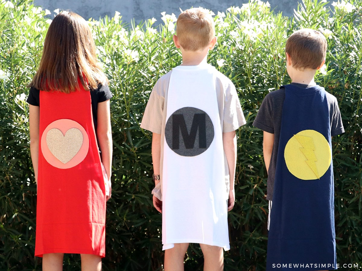 three kids standing outside with their backs to the camera showing off their superhero capes