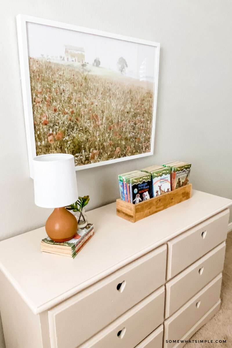 pink dresser with a lamp and books on top and a framed print hanging on the wall