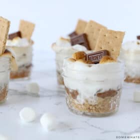 smores in small jars on a white counter with marshmallows sprinkled around