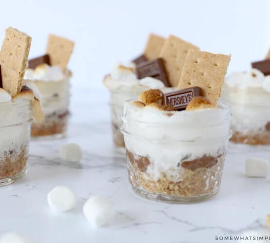 smores in small jars on a white counter with marshmallows sprinkled around