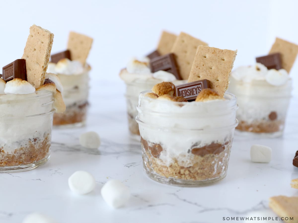 smores in small jars on a white counter with marshmallows sprinkled around