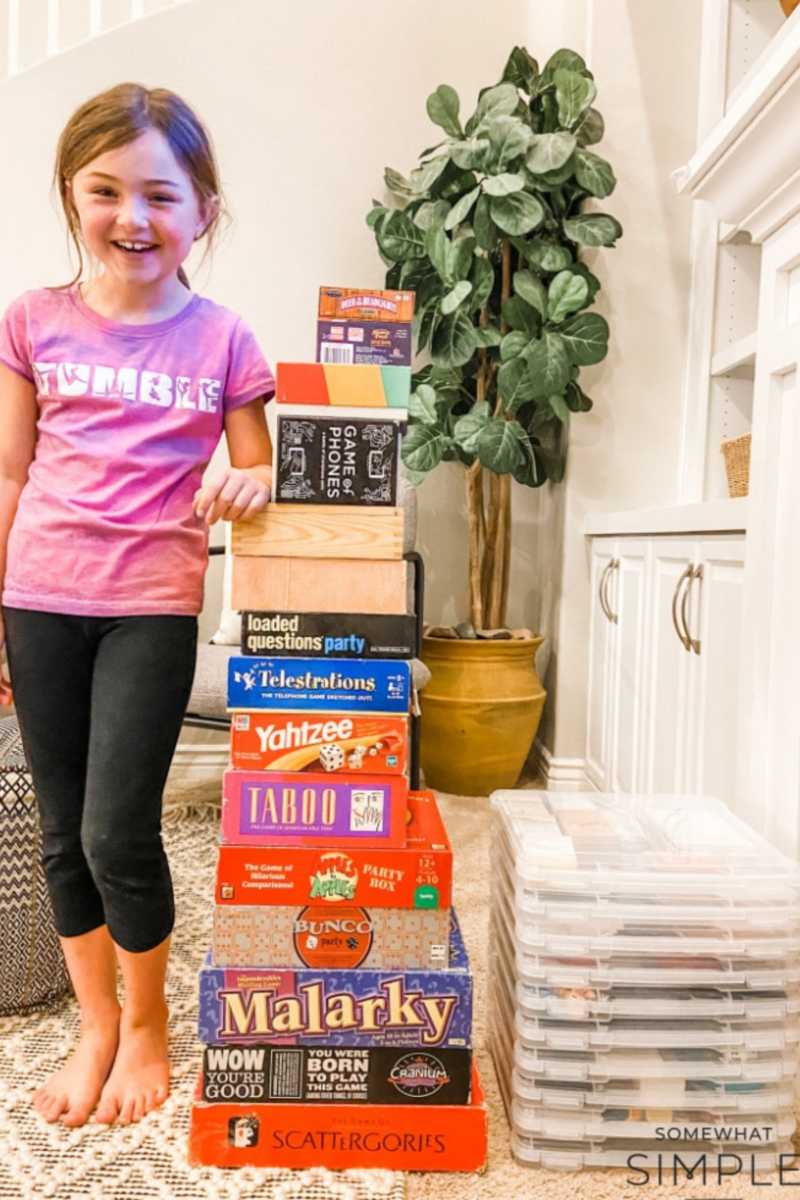 a little girls standing next to a tower of board games to illustrate how much space they take up