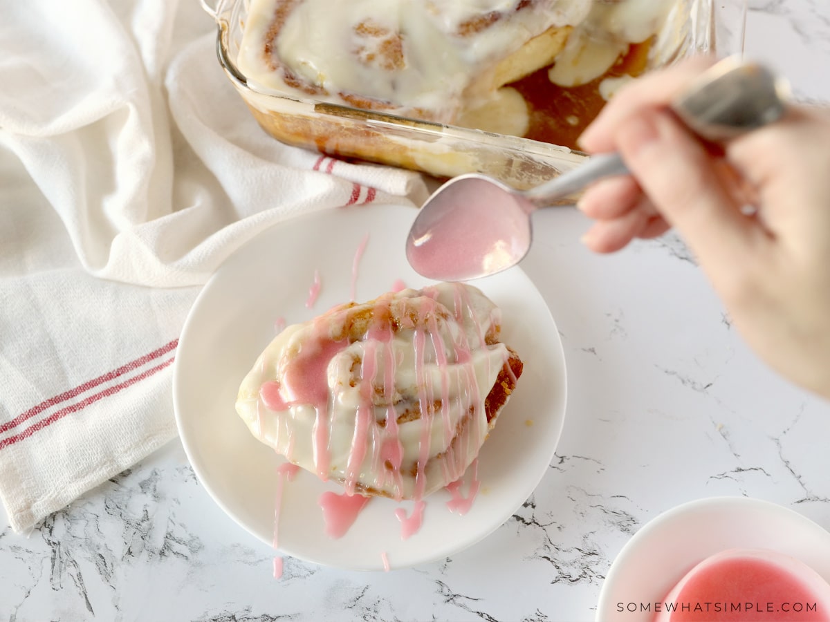 drizzling pink frosting over the top of valentine cinnamon rolls